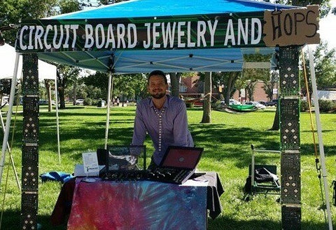 Drew at an out door market smiliing while standing his his TechWears Booth. The sign now reads "Circuit Board Jewlery and Hops" The "Hops" portion of the sign is wrote in sharpie on cardboard and clipped to the sign in a makeshift type way.