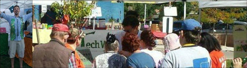 Drew stands proud wearing a vest Techwears has made from upcycled circuit board. Drew is surrounded by customers at a market. 